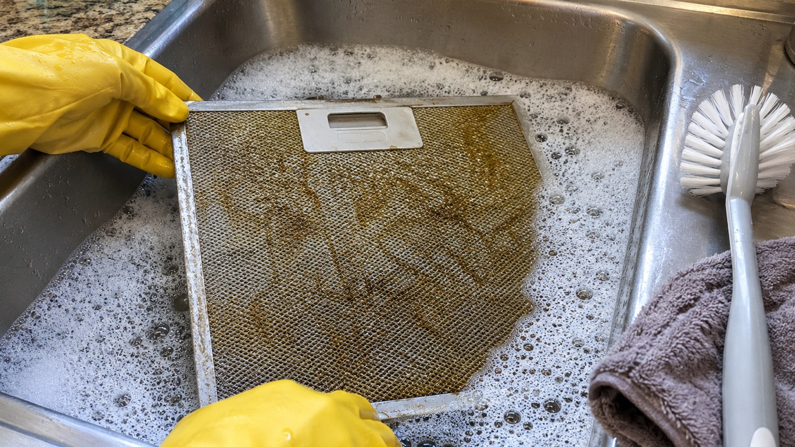 Metal range hood filter soaking in soapy water in a kitchen sink with a soft brush and towel nearby.
