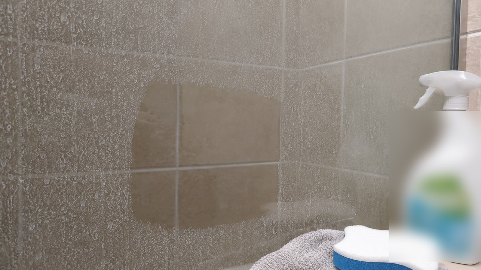 Apartment shower wall with light soap scum, a non-scratch sponge, towel, and mild bathroom cleaning supplies.